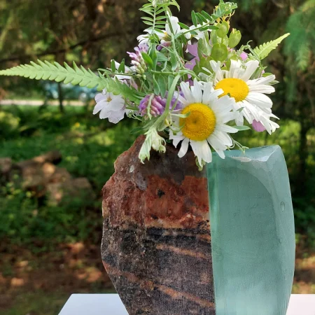 The artist’s work displayed in the outdoors on a white plinth. A vase that is half glass half stone holds fresh cut wildflowers.