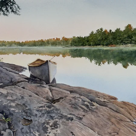 A landscape scene; a canoe and paddle are set up on a rocky shoreline on a calm lake. There is a forest, mainly of pine trees in the distance, which is reflected in the water.