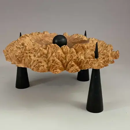 A highly textured piece of wood, with a black ball sitting in the centre, is supported off the ground by three black cones that penetrate the wood on three sides.