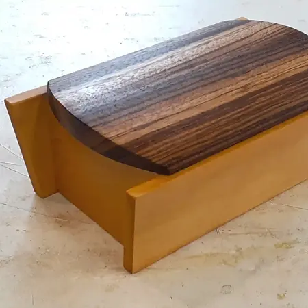 A closed wooden box; the top is made of a dark wood with a distinctive wood grain. The base is made from a lighter wood that forms a square box with the horizonal sides extending beyond the vertical sides.