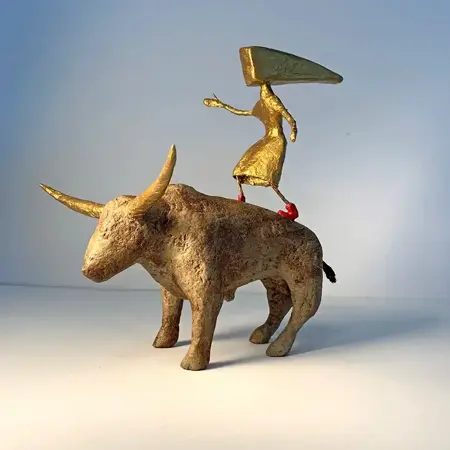 A gold human figure with an irregular shaped head, wearing red shoes stands on the back of an gold bull with horns.