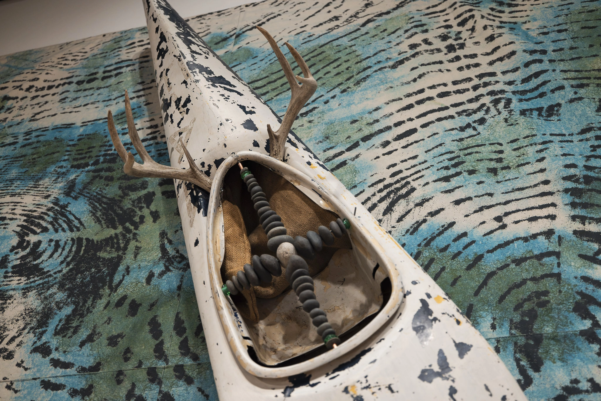 A weathered white kayak with dark splotches, adorned with antlers and beads, is set on a textured blue and green patterned background.
