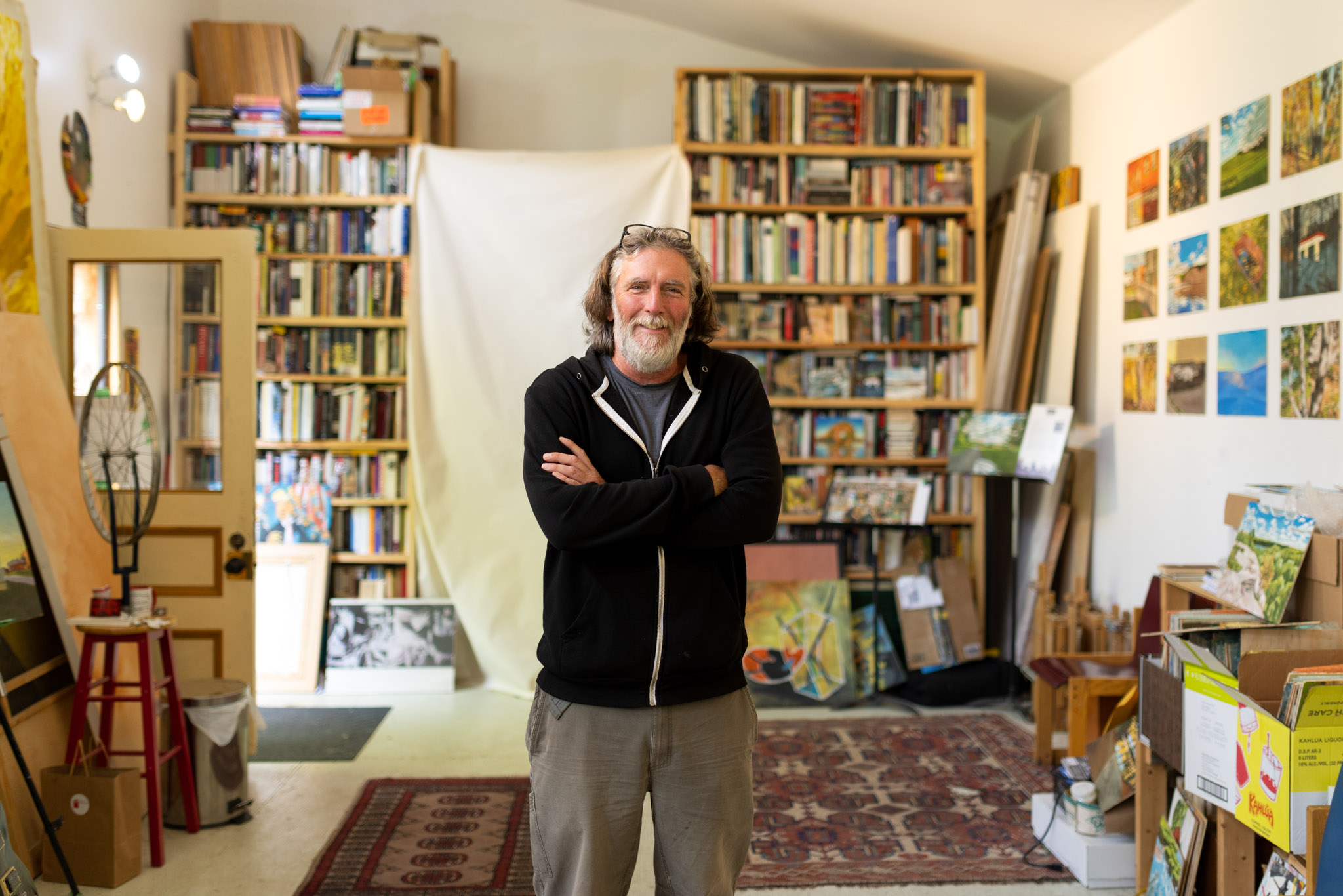 John Climenhage stands in his studio smiling with his arms corssed. Behind him are several bookshelves filled with books, and on the right is a gallery wall full of brightly coloured paintings 