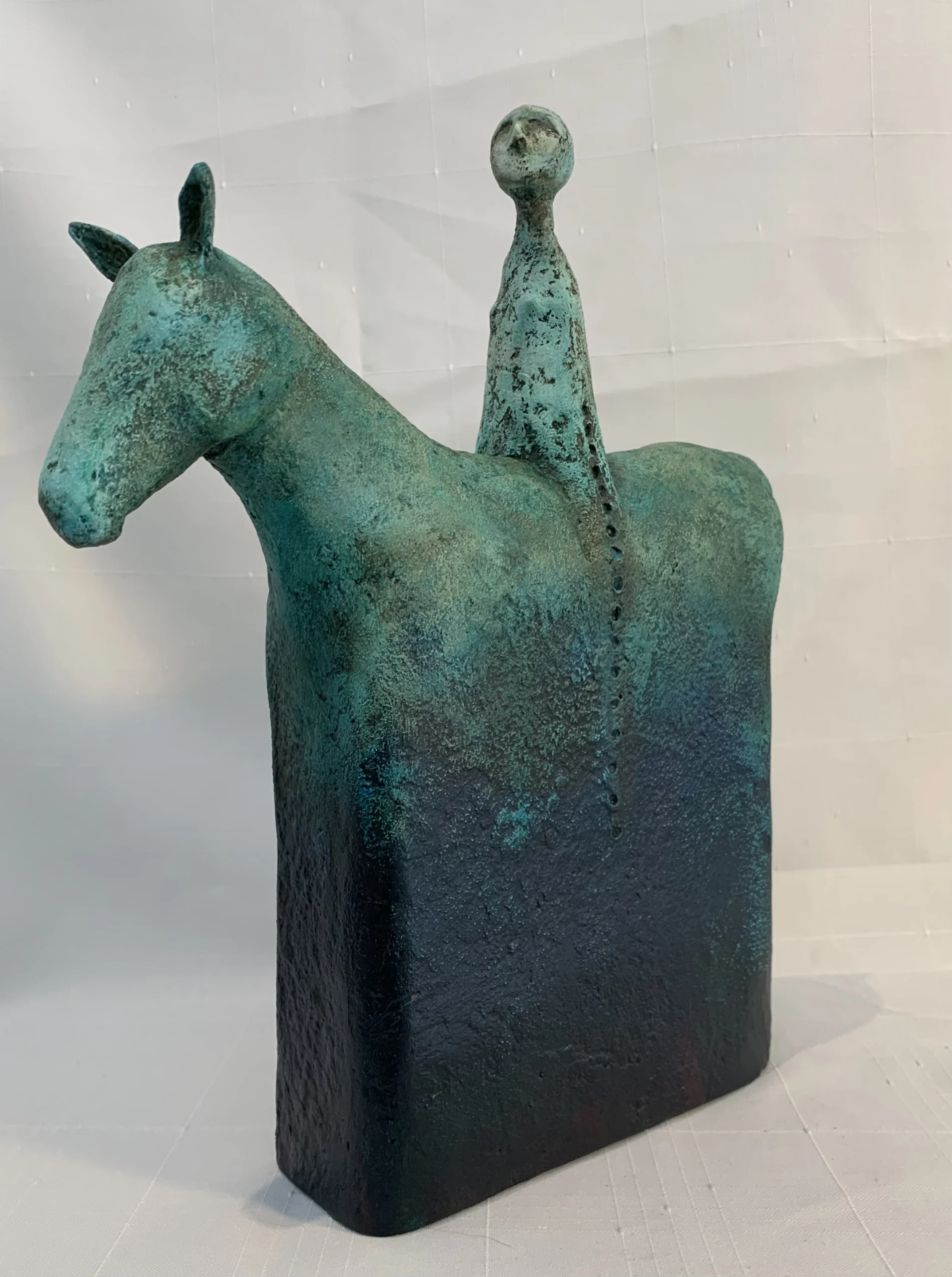Annette Vlieg’s sculpture of an abstract sculpture portrays a simplified horse with a tall, elongated rider figure. The sculpture is painted in a gradient starting as turquoise at the top and moving to black at the bottom.