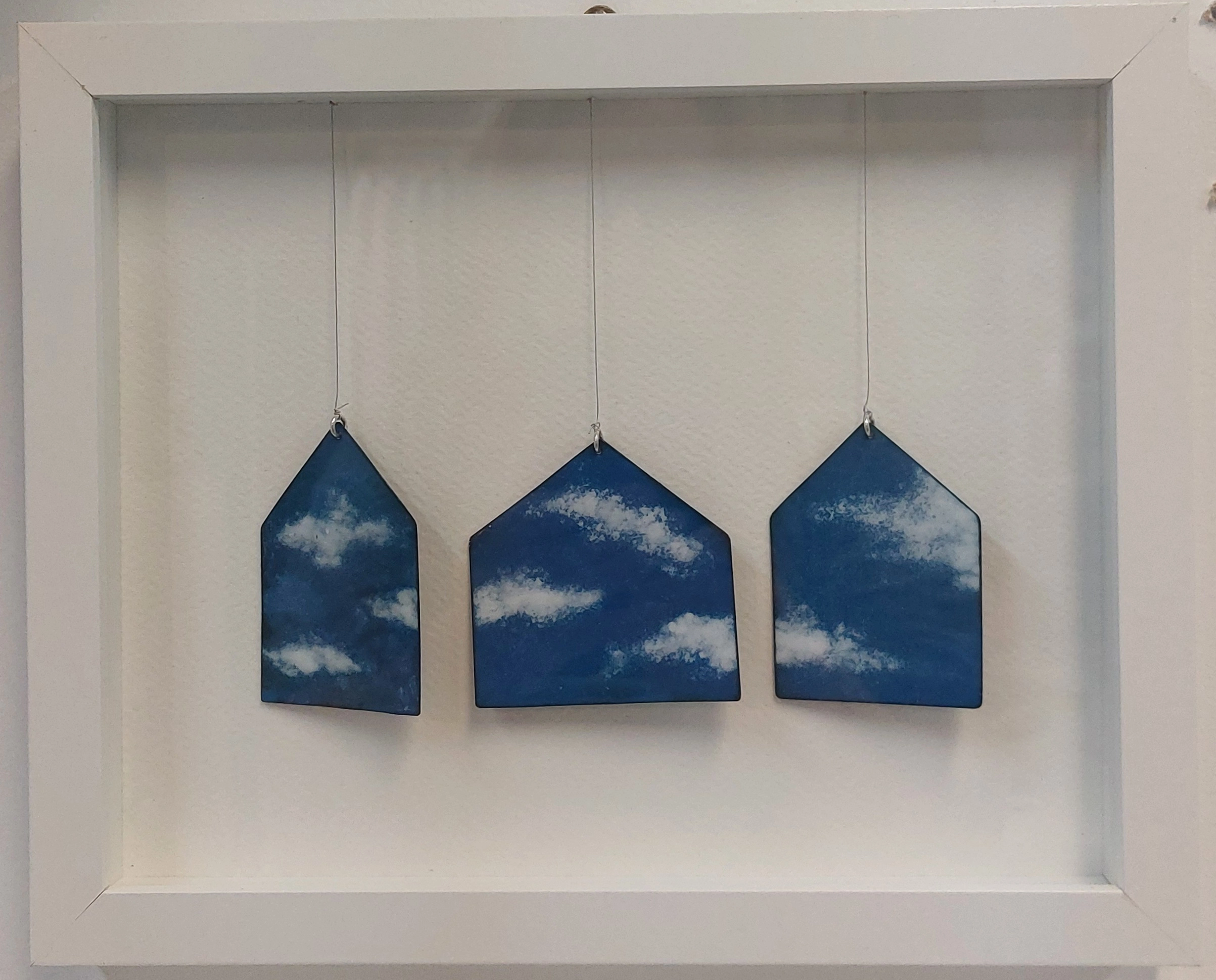 Photograph of three house-shaped pendants hanging inside a white frame, each painted with a blue sky and white clouds. Created by Christy Haldane titled “Dreaming of Home”.