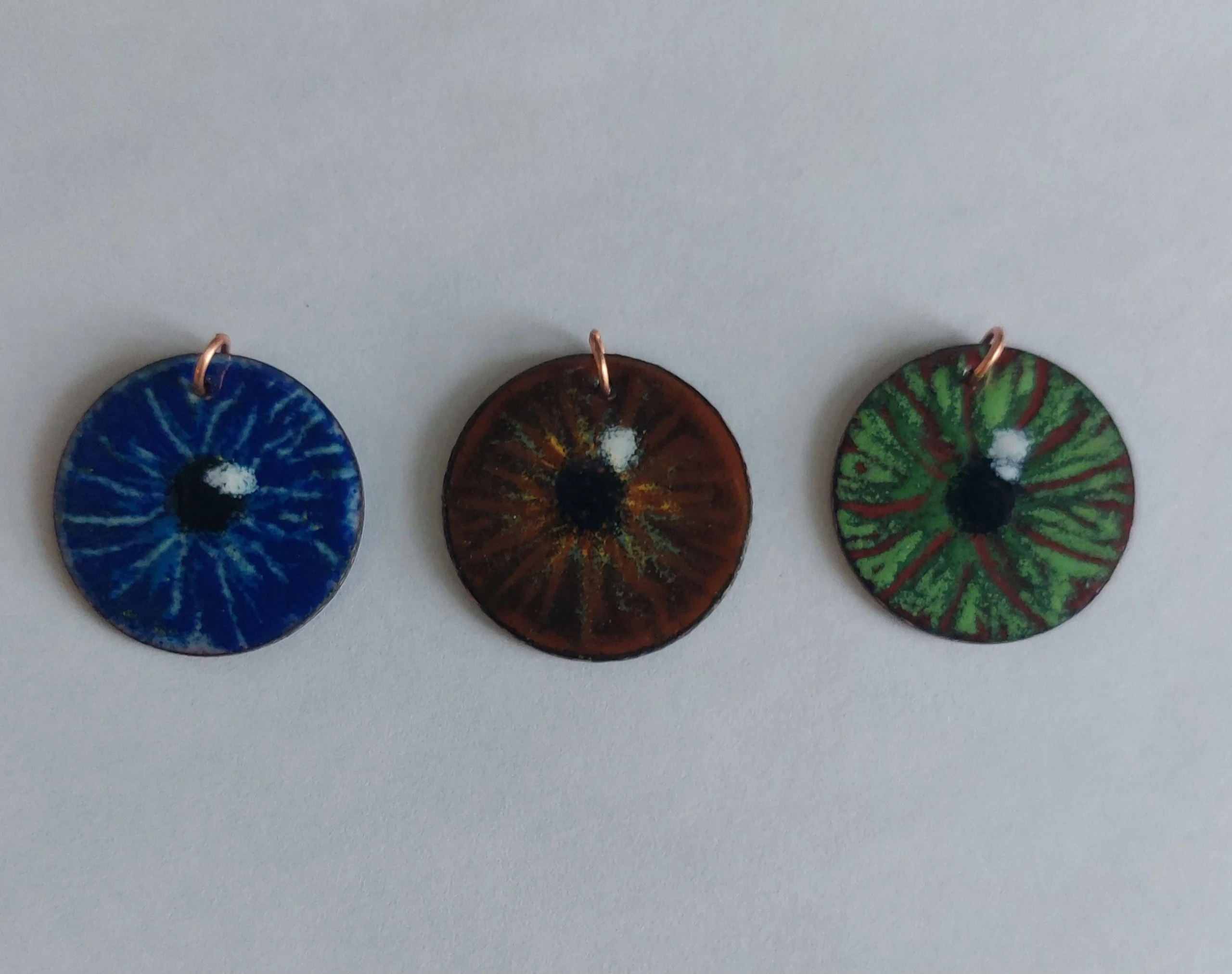 Photograph of three circular pendants created by Christy Haldane that resemble human irises in blue, brown, and green colors. Each pendant features detailed radial patterns and a small metal loop at the top for attachment. Titled “Eyes”.