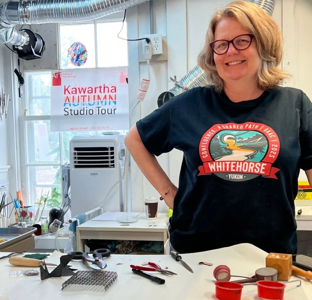 Photograph of Christy Haldane standing in their art studio with various tools and materials on a worktable, including wire mesh, pliers, and tape.