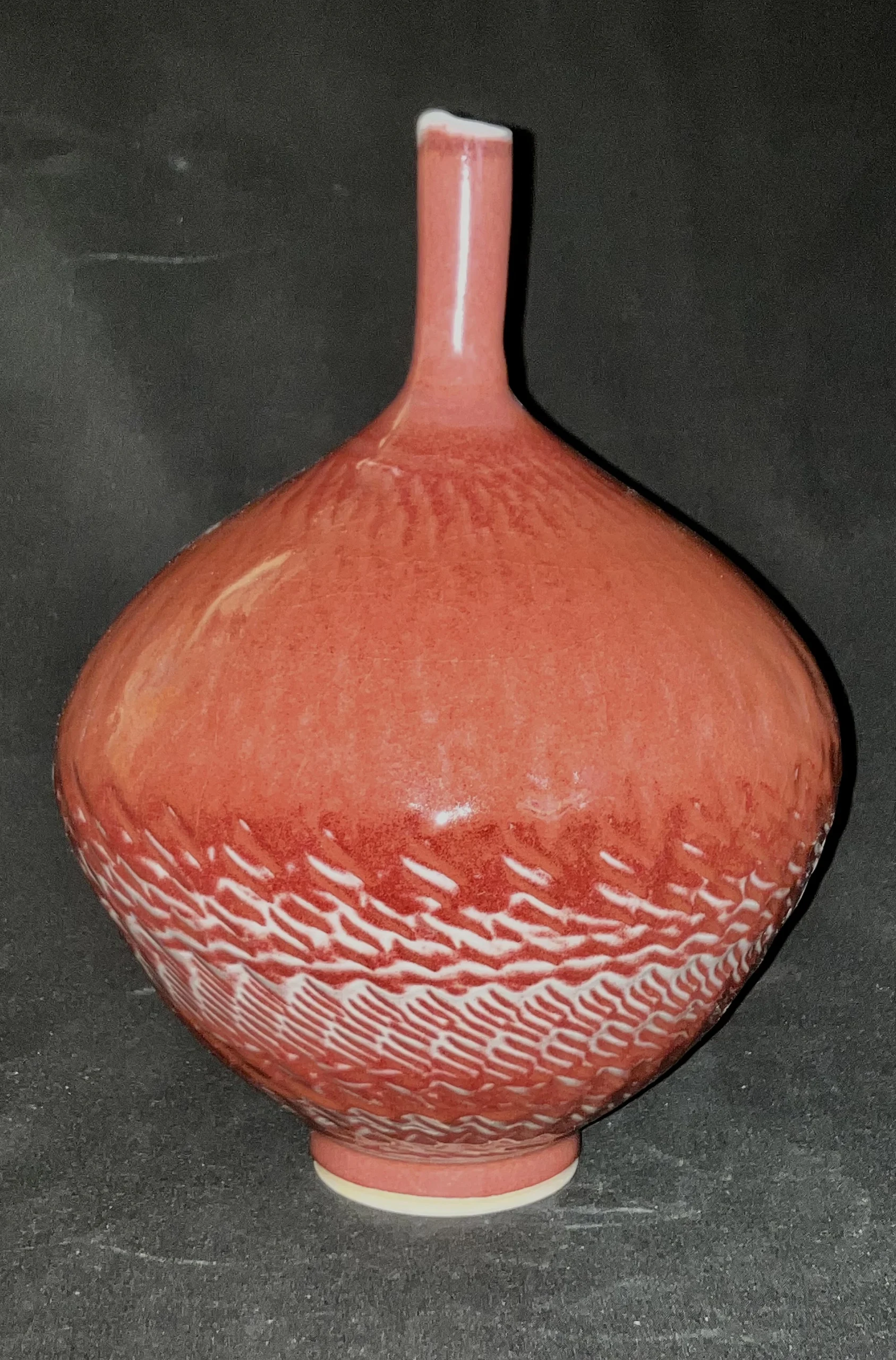 Photograph of a porcelain vase with a bulbous body and narrow neck, featuring a textured surface and a reddish-orange glaze with white accents near the base. Created by Don Clark titled “Long Necked Bottle”.