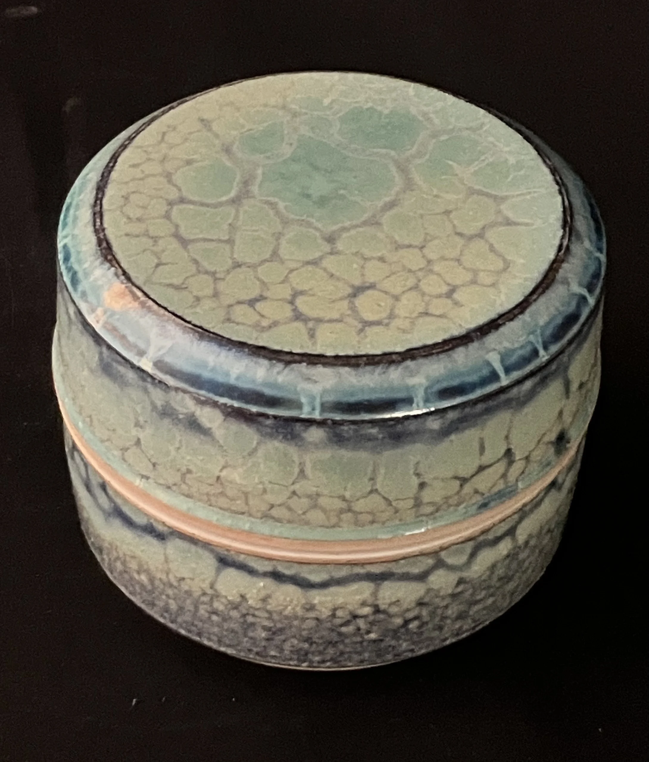 Photograph of a small round porcelain jar with a lid, featuring a crackled glaze pattern in shades of green, blue, and beige. Created by Don Clark titled “Small Lidded Jar”.