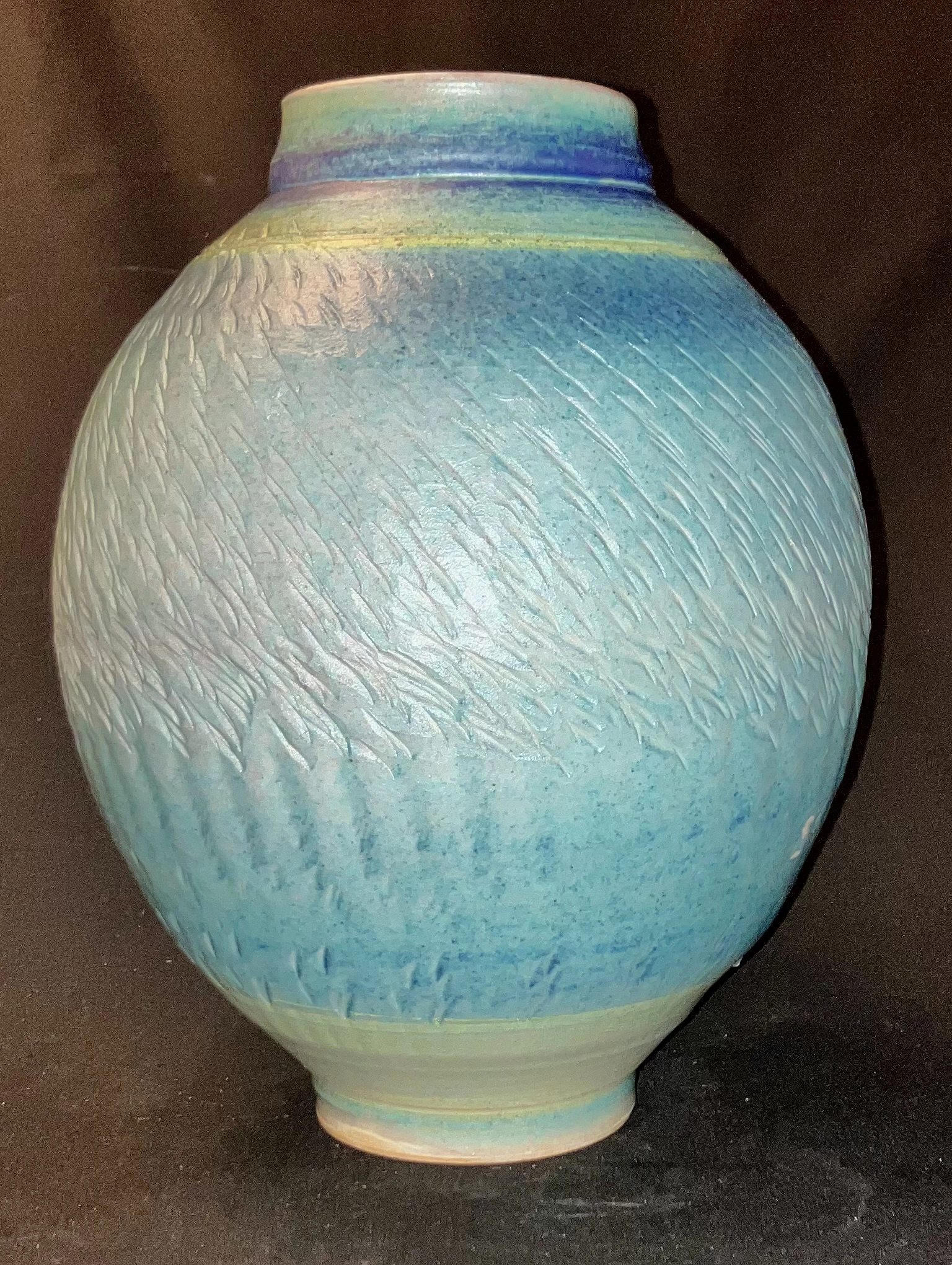 Photograph of a rounded ceramic vase with a narrow neck, featuring textured surface patterns and a gradient glaze transitioning from light blue to darker blue and hints of yellow near the top. Created by Don Clark titled “Vase, Copper Blue Matt Glaze”