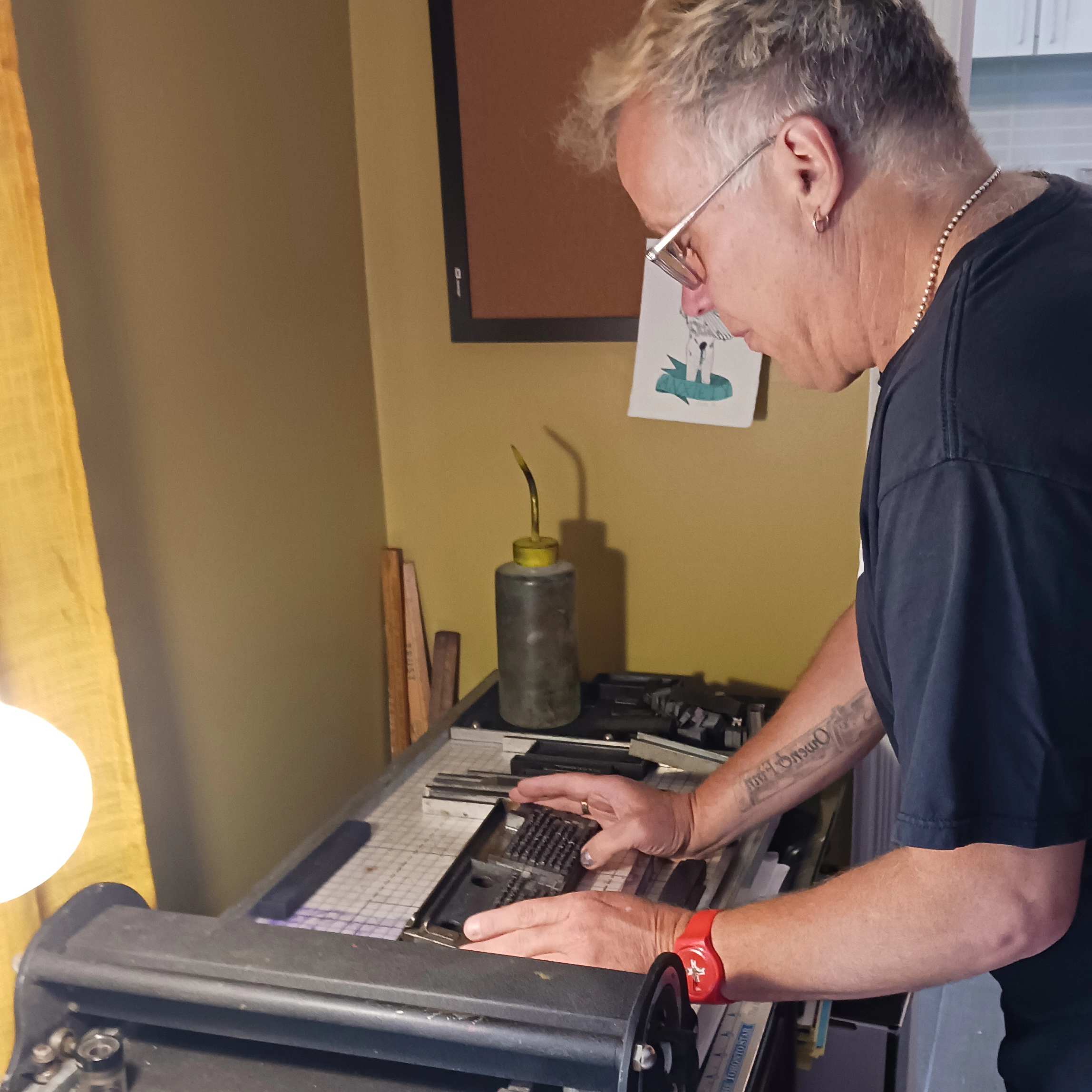 Photograph of Jeffrey Macklin operating a letterpress machine in their studio setting, focusing on hands adjusting metal type on the press bed.