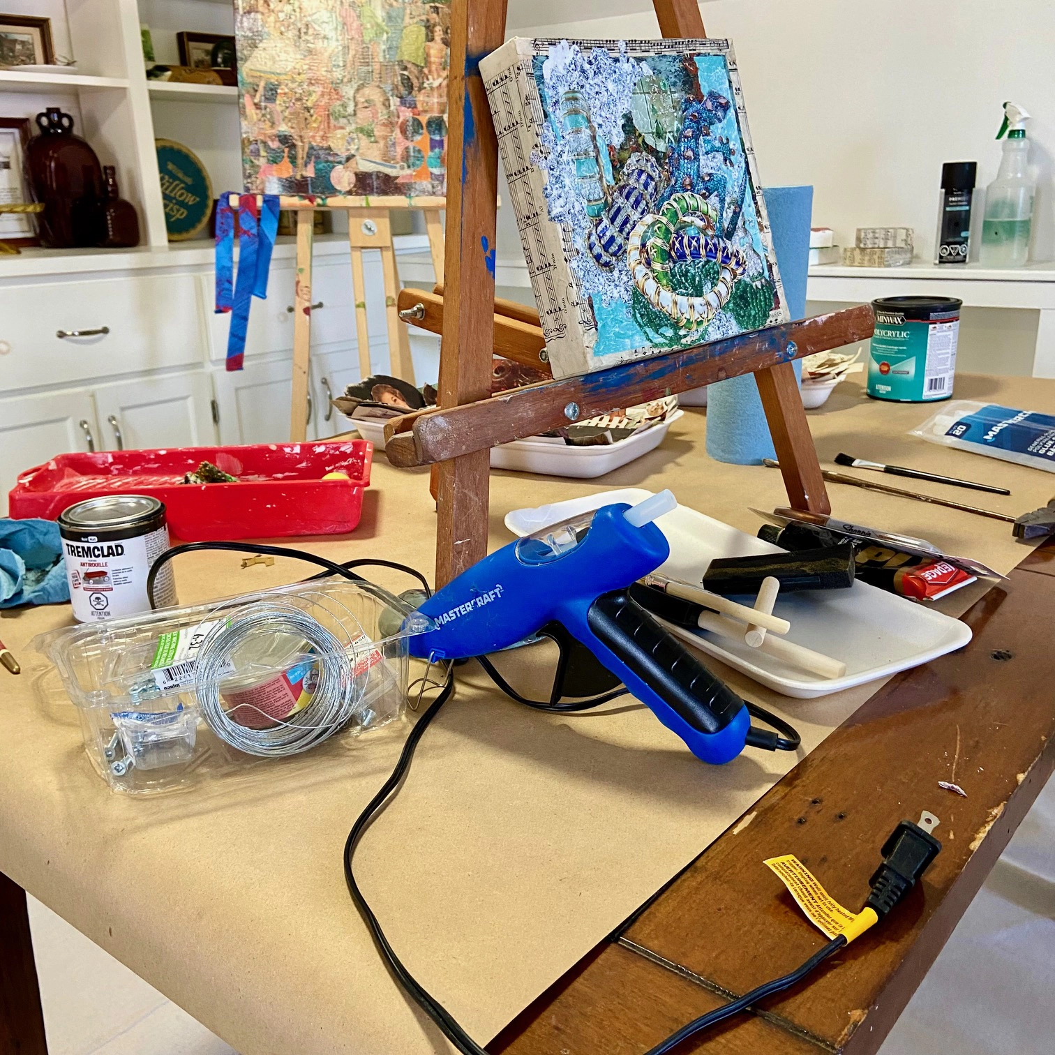 Photograph of Kay Gregg’s studio featuring a wooden easel holding a small canvas with a colorful, abstract painting. Surrounding the easel are various art supplies.