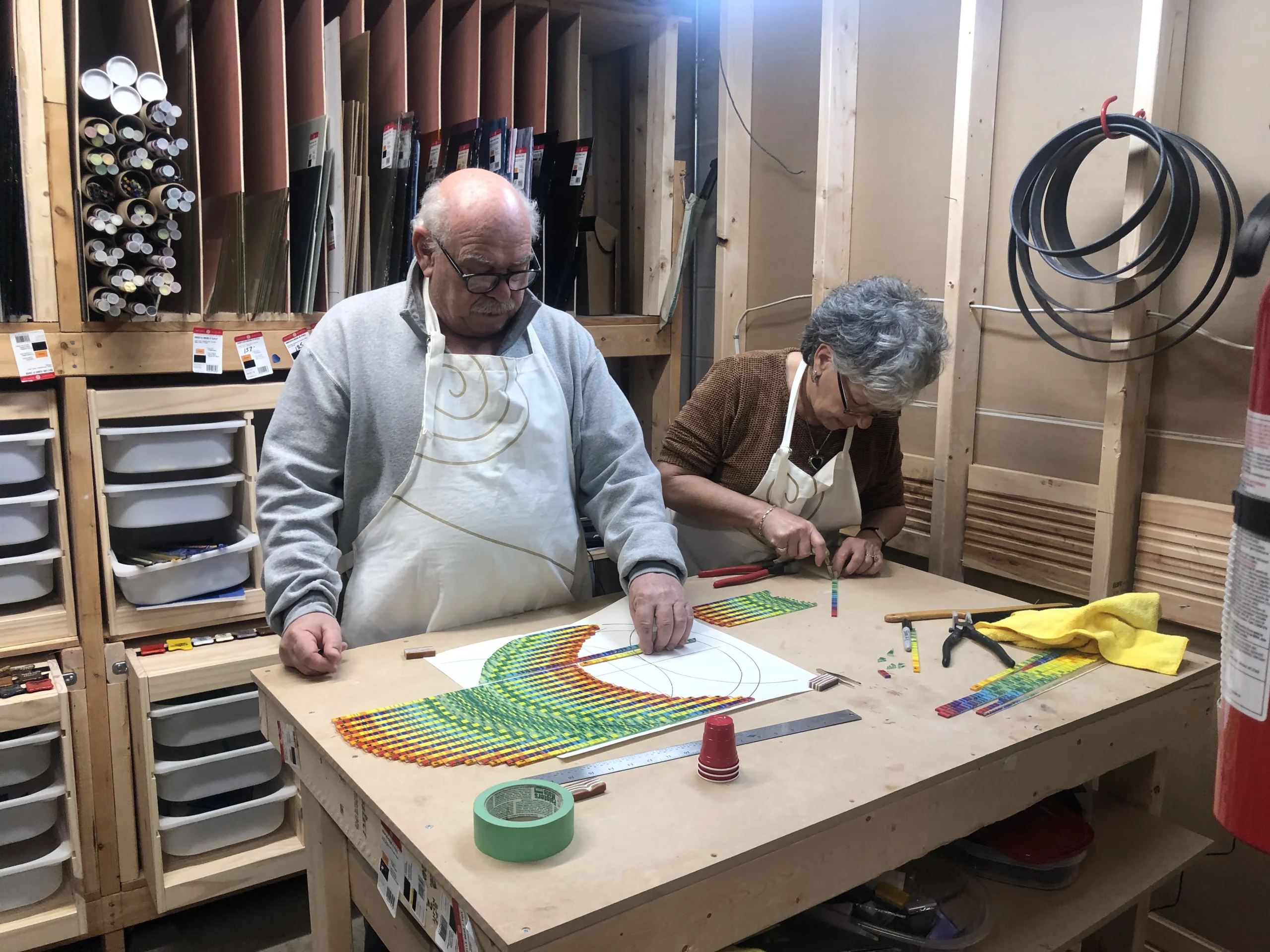 Photograph shows Lesley and Al Kroach working on a large, colorful mosaic or tile art project on a wooden table in their studio.
