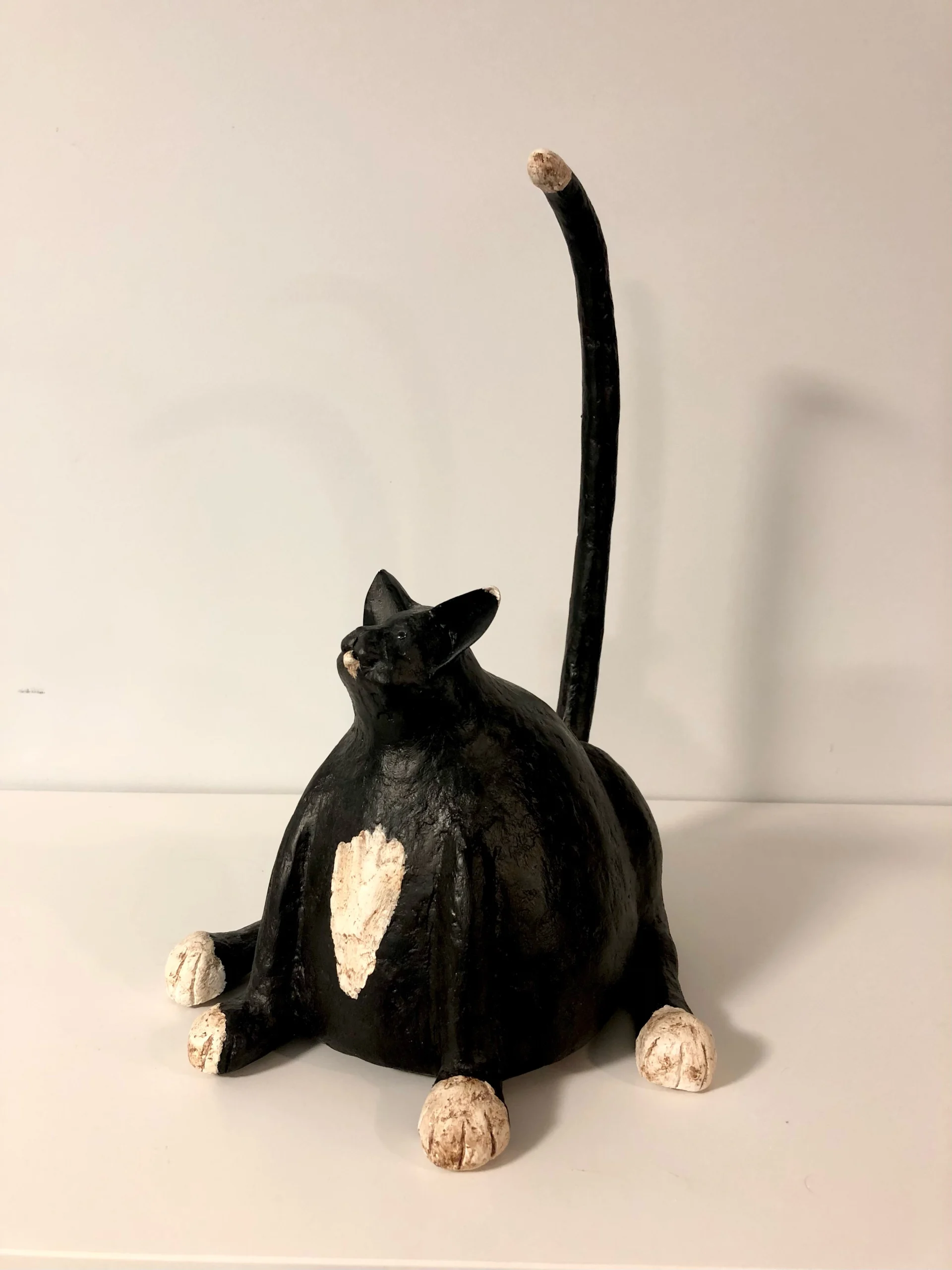 Sculpture of a black cat with a white patch on its chest and white paws, sitting with an exaggeratedly long, upright tail. The cat has a rounded body and a slightly tilted head, placed against a plain light background. Created by Mark Hinan titled “Boots”