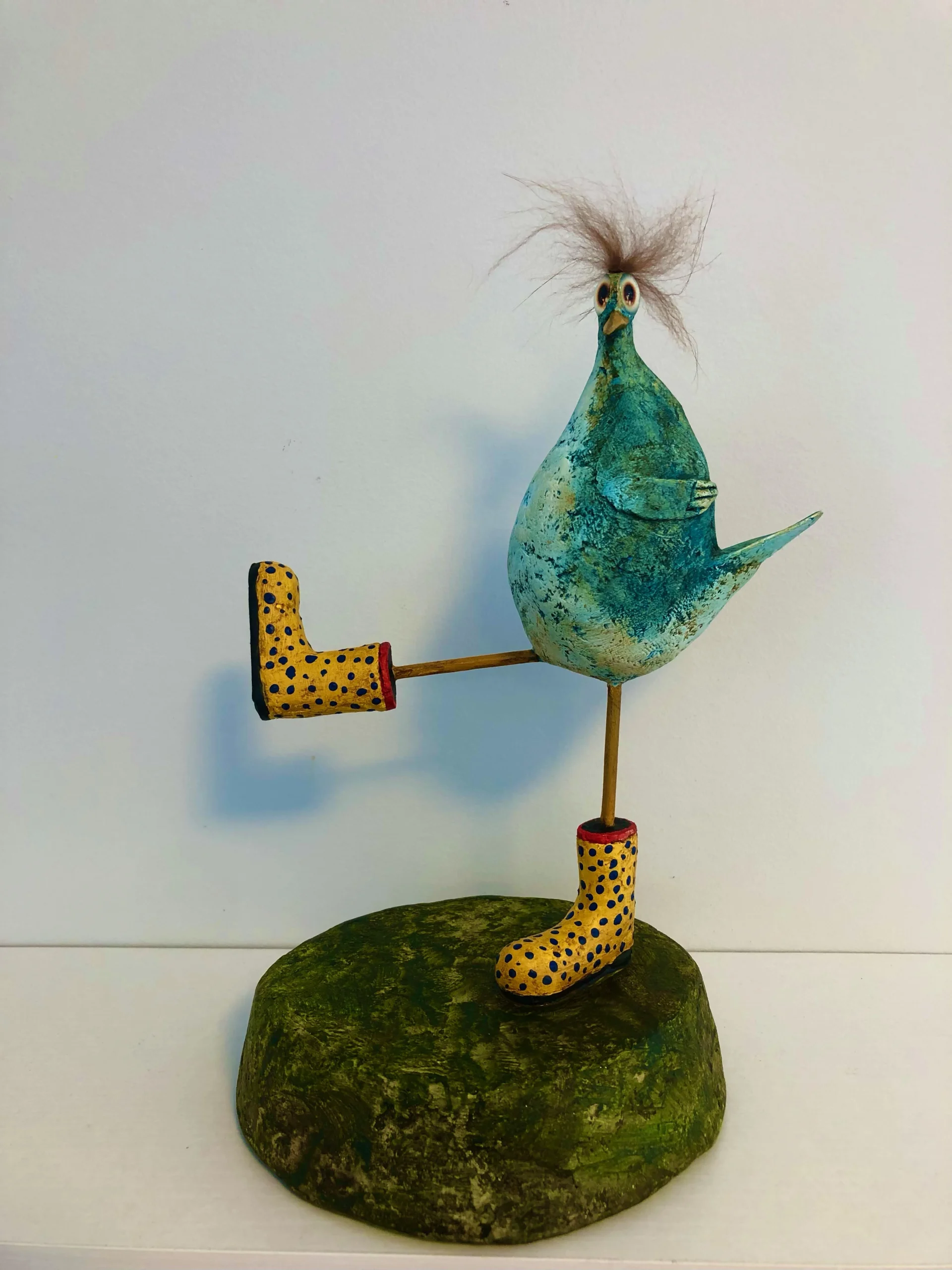 Photograph of bird sculpture standing on one leg atop a green, textured base. The bird has a blue-green body, wild brown hair, wide eyes, and wears yellow boots with black spots and red trim. Created by Mark Hinan titled “Matilda Merryweather”.