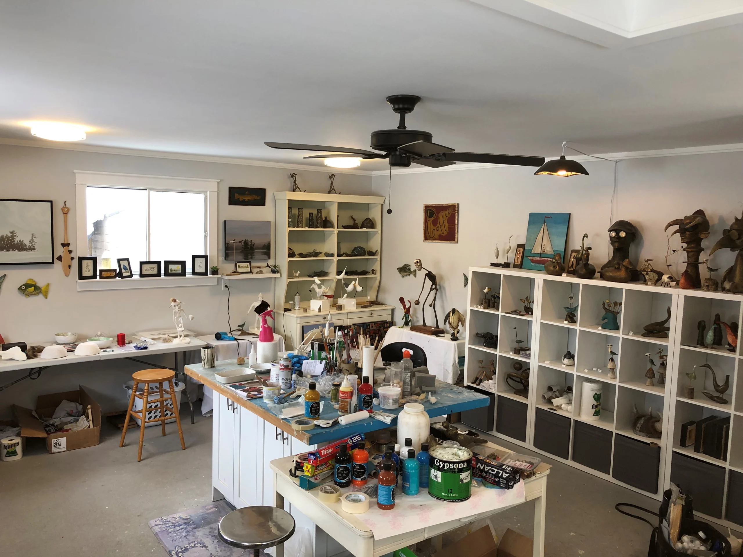 Photograph of Mark Hinan’s studio featuring a central worktable with paint bottles, brushes, and art supplies, surrounded by shelves and cubbies filled with sculptures and decorative objects.