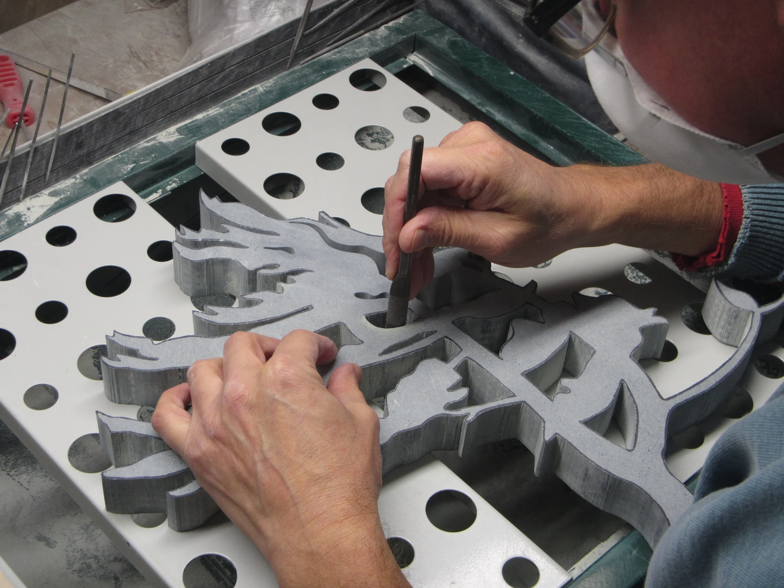 Photograph of Nick Leniuk using a tool to carve or refine a detailed, multi-layered sculpture made of gray material on a perforated work surface.