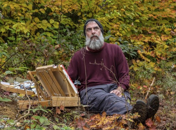 Photograph of Paul Nabuurs sitting on the ground outdoors surrounded by autumn foliage, engaged in painting with an easel and art supplies nearby.