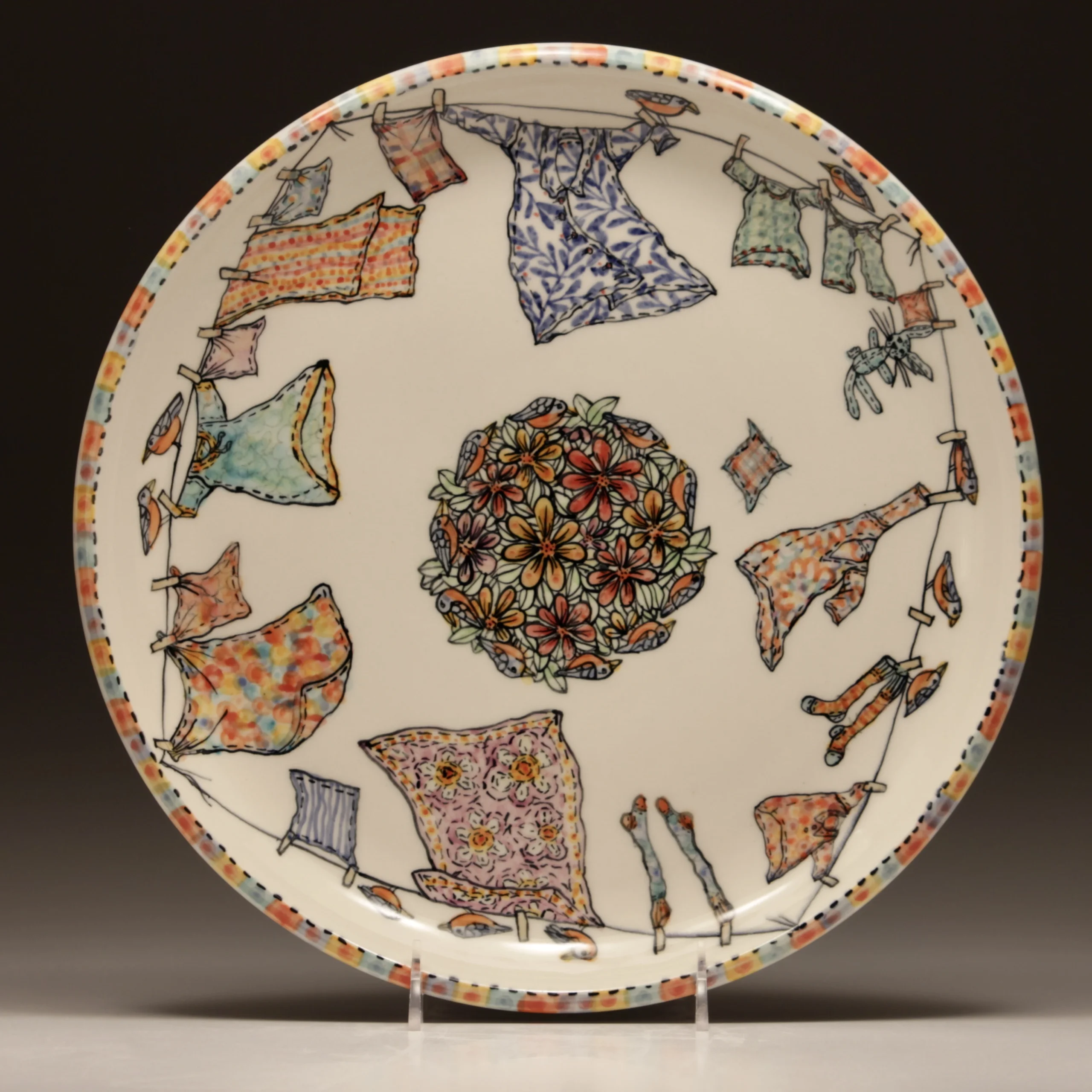 Decorative Porcelain plate by Thomas Aitken & Kate Hyde features hand-painted clothes hanging on a clothesline around a central floral pattern with multicolored flowers. Plate rim has a dotted border in orange, blue, and black.