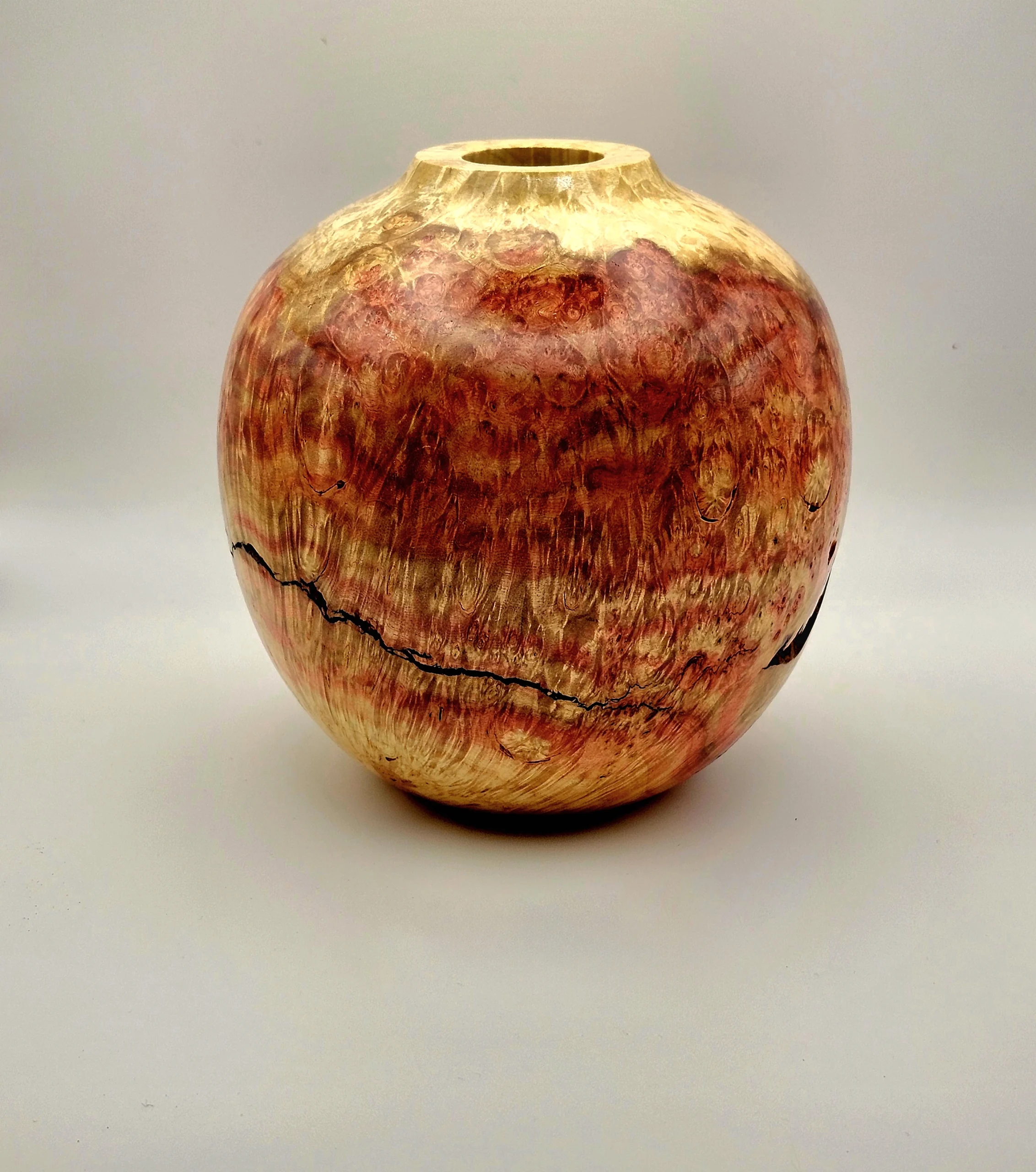 Photograph of “Boxelder Burl” a handcrafted wooden object with a rounded shape and narrow opening at top. Object features rich reddish-brown and golden hues with natural grain patterns and a prominent black crack running horizontally around its midsection. Created by Vince Way-Nee