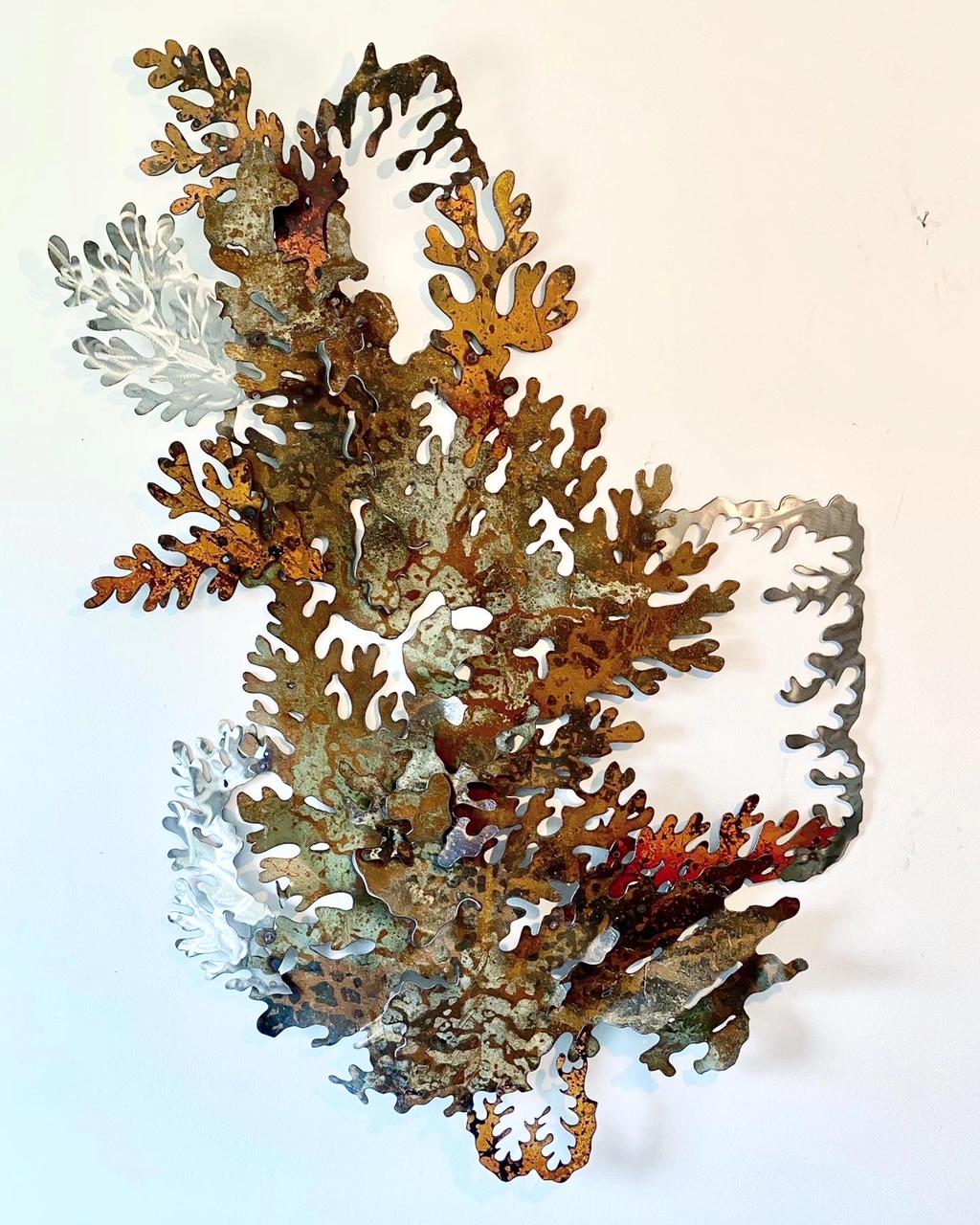 A photograph of a metal sculpture created by Garrett Gilbart resembling layered mosses with intricate cut-out patterns. The sculpture features a mix of rusted brown, green, and silver tones. Titled “Lichen Composition”.